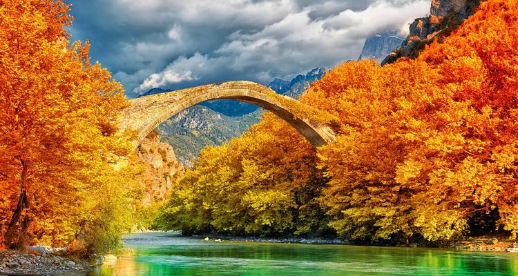 Un élégant pont en pierre en arc enjambe une rivière turquoise encadrée par un feuillage automnal flamboyant et des montagnes imposantes.