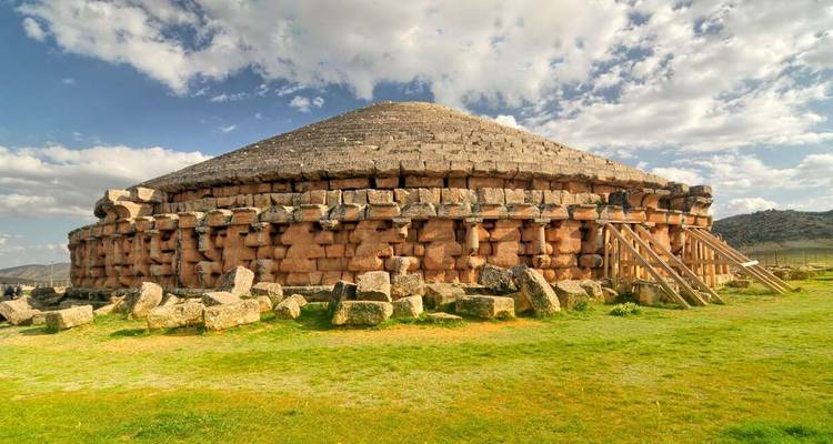Circular stone mausoleum built of large granite blocks set on grassy plains under a cloudy sky