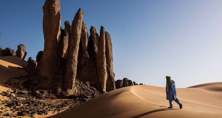 Lone traveller in blue robe walking across wind-rippled dune toward towering stone spires