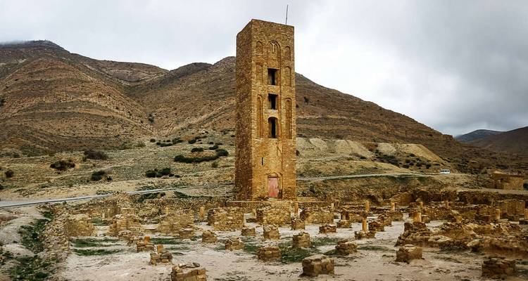 Tall stone tower rising above scattered ruins in a rugged mountain valley under grey skies