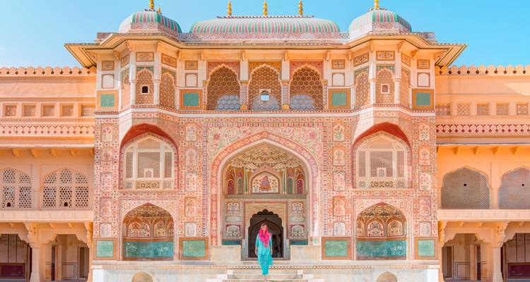 Eine Frau in farbenfroher traditioneller Kleidung steht vor dem reich verzierten Eingangstor des Amber Fort und präsentiert die aufwendige rajasthanische Architektur unter einem klaren blauen Himmel.