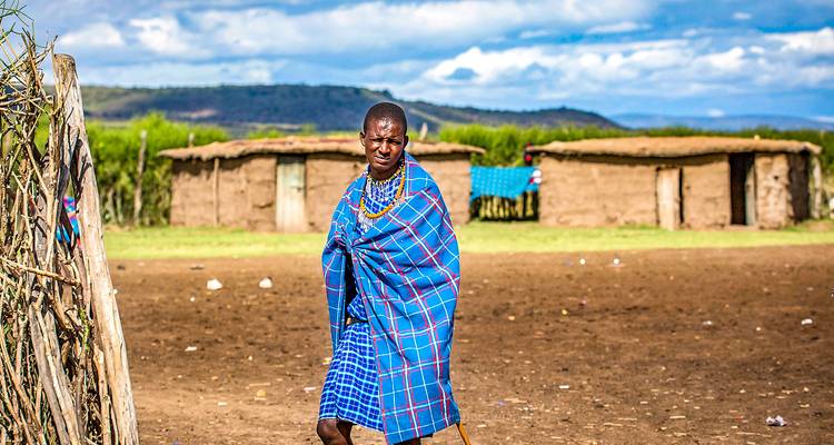 Maasai man gewikkeld in blauwe shuka staat voor traditionele leemhutten op open vlakte