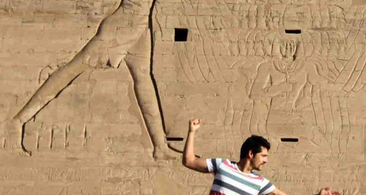 Traveller strikes a playful pose beside carved reliefs on an ancient sandstone temple wall
