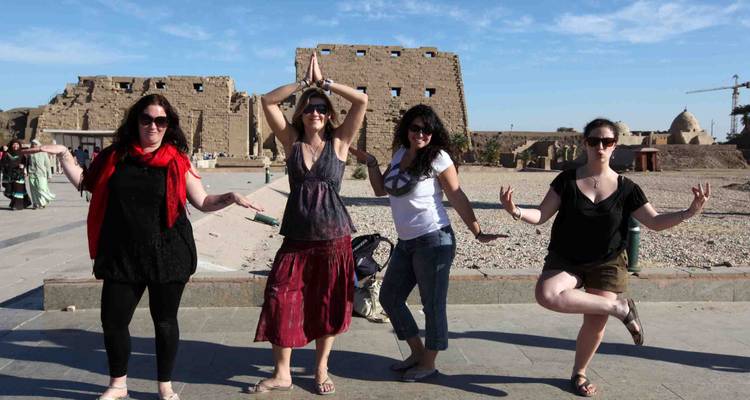Four female travellers strike playful poses in front of ancient sandstone temple ruins under a clear blue sky.