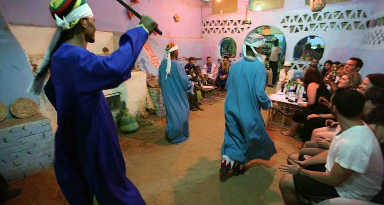 Traditional dancers in blue robes perform with sticks before an audience inside a rustic, sand-floored room.