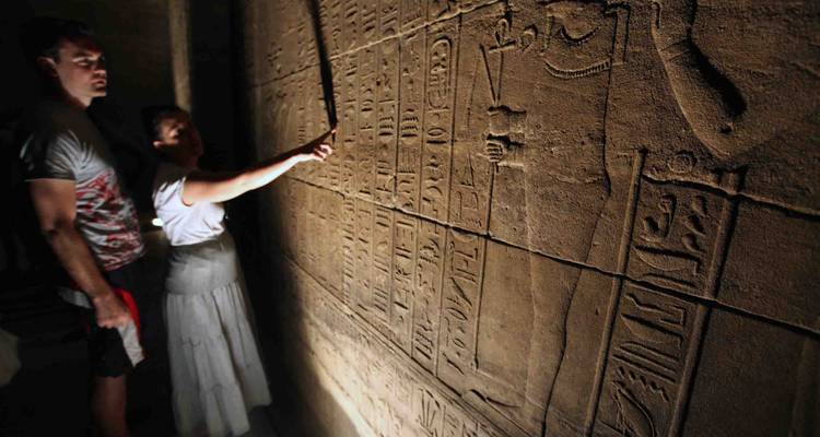 Two visitors study hieroglyphic carvings on a sandstone wall inside an ancient Egyptian temple passage.