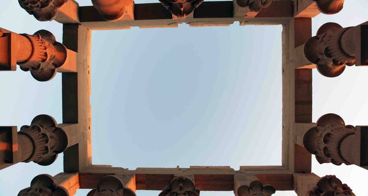 Upward view through a square opening framed by ancient stone columns glowing in warm light against a clear sky.