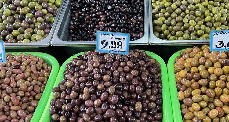 Variétés d'olives empilées en hauteur sur un étal de marché grec avec panneau de prix.