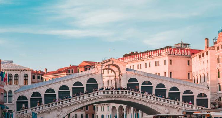 Le pont emblématique du Rialto enjambe le Grand Canal de Venise bordé de bâtiments pastel sous un ciel dégagé.