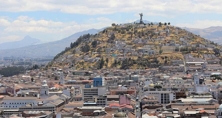 Breed stadsgezicht van Quito's daken die zich uitstrekken richting de El Panecillo heuvel bekroond door het Mariabeeld, met verre Andestoppen.