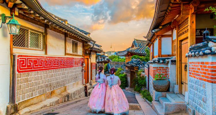Deux femmes en hanbok rose marchent dans une ruelle colorée du village de Bukchon Hanok au coucher du soleil avec la tour N Seoul en vue.
