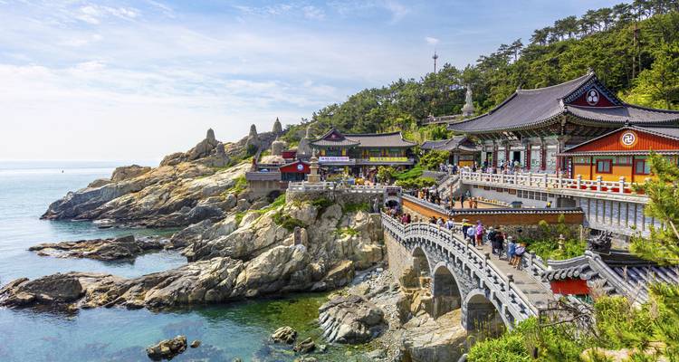 Temple Haedong Yonggungsa perché sur des falaises rocheuses au-dessus de la mer avec des visiteurs traversant un pont de pierre.
