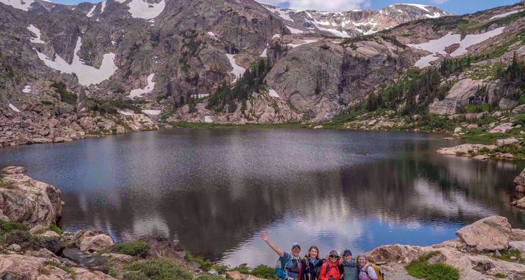 Quatre randonneurs souriants posant près d'un lac alpin immaculé reflétant des montagnes escarpées striées de neige.