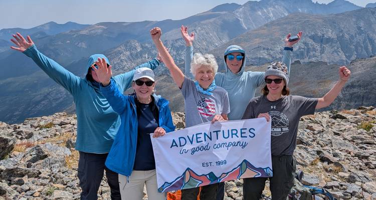 Cinq randonneurs jubilants debout sur un sommet rocheux tenant une bannière avec des montagnes spectaculaires en arrière-plan.