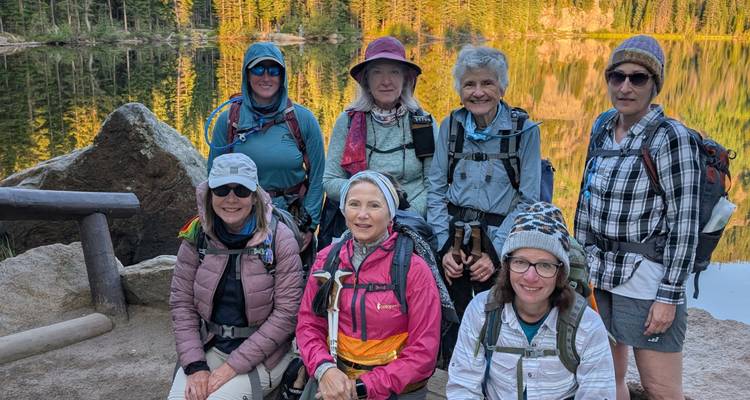 Sept femmes randonneuses posant près d'un lac tranquille avec la forêt et les reflets dorés du soleil.