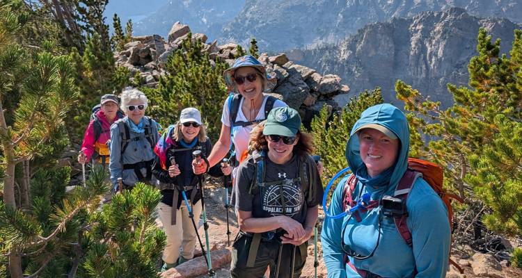 Ligne de randonneuses souriantes le long d'une crête rocheuse étroite encadrée par des pins et des falaises imposantes.