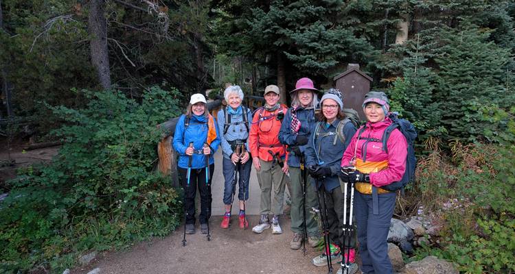 Six randonneurs debout sur un sentier forestier ombragé entouré de conifères denses.