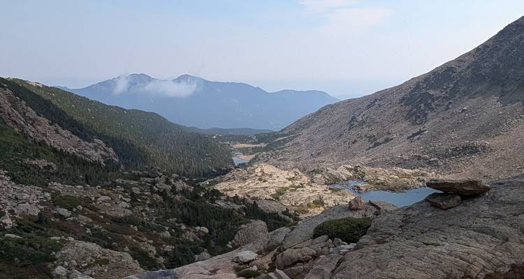 Vaste vallée de montagne avec des lacs alpins épars and des crêtes rocheuses sous une lumière du jour brumeuse.