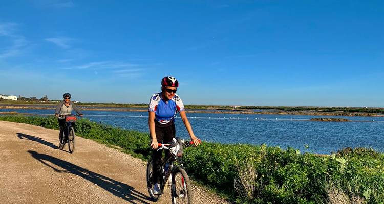 Deux cyclistes pédalent le long d'un chemin de terre au bord d'un lagon bleu parsemé de flamants roses au loin.