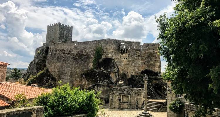 Eine robuste mittelalterliche Steinburg erhebt sich gegen einen teilweise bewölkten Himmel, umgeben von Grün und Ziegeldächern