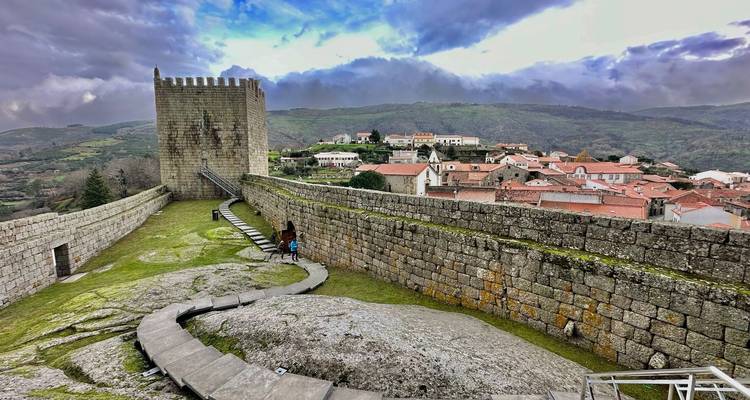Besucher erkunden die Wälle und den Turm einer antiken Hügelfestung mit Blick auf ein rotgedecktes Dorf