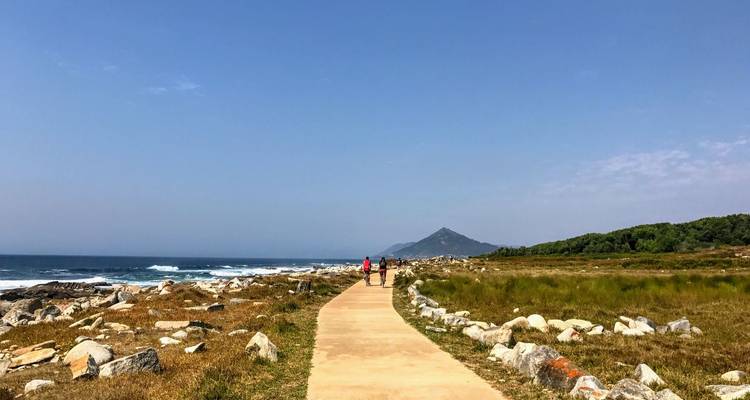 Dos personas caminan por un sendero pavimentado junto al mar flanqueado por rocas y vegetación baja con vista a montañas distantes