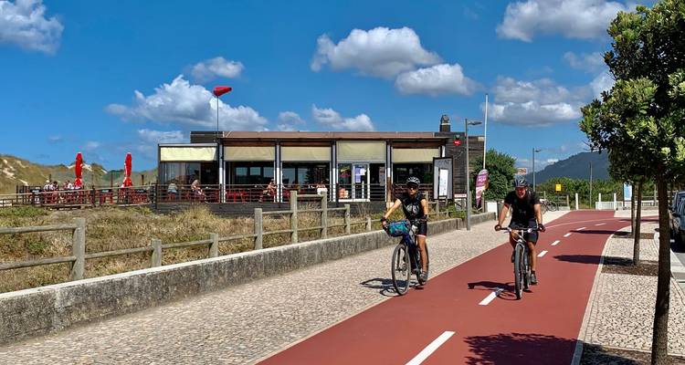 Los ciclistas cruzan por un carril bici rojo pasando por un café frente a la playa bajo pequeñas nubes esponjosas