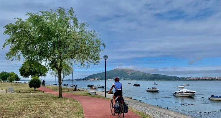 Un cyclotouriste pédale le long d'un sentier au bord de l'eau avec des bateaux amarrés et une colline verte basse au-delà