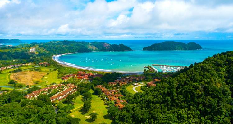 Prise de vue panoramique par drone d'une baie en forme de croissant, d'un port de plaisance et de collines luxuriantes rencontrant les eaux turquoise du Pacifique.