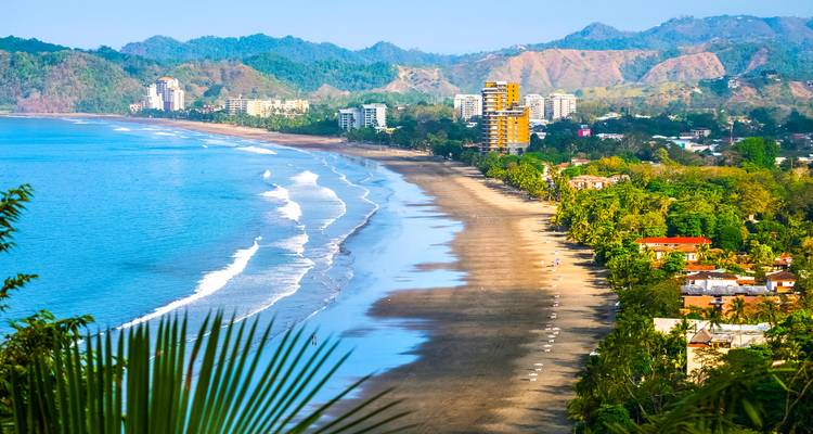 Longue étendue de la plage de Jaco bordée de palmiers et de complexes hôteliers au loin près d'une mer bleu vif.