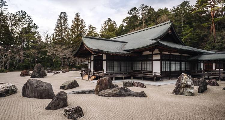 Jardín zen de rocas con arena meticulosamente rastrillada y un salón tradicional de madera rodeado de árboles altos.