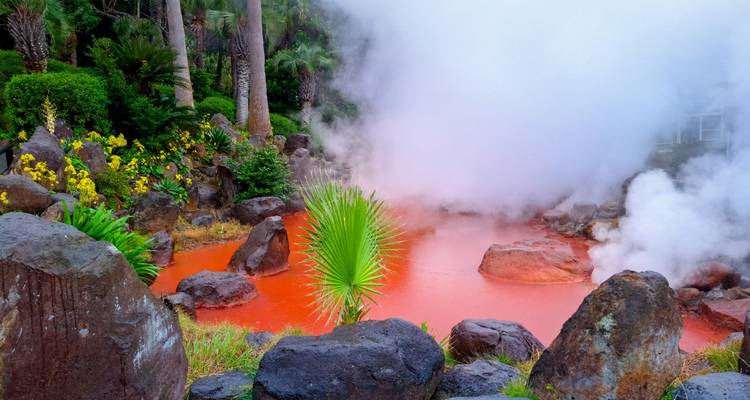 Bassin de source chaude orange vif émettant de la vapeur au milieu de plantes tropicales et de roches volcaniques.