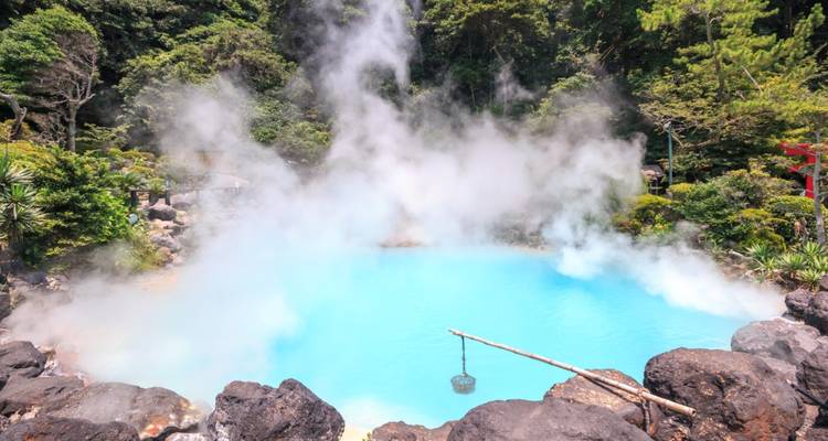 Piscine géothermale bleu laiteux entourée de rochers noirs et d'une forêt luxuriante, avec une vapeur épaisse qui s'élève.