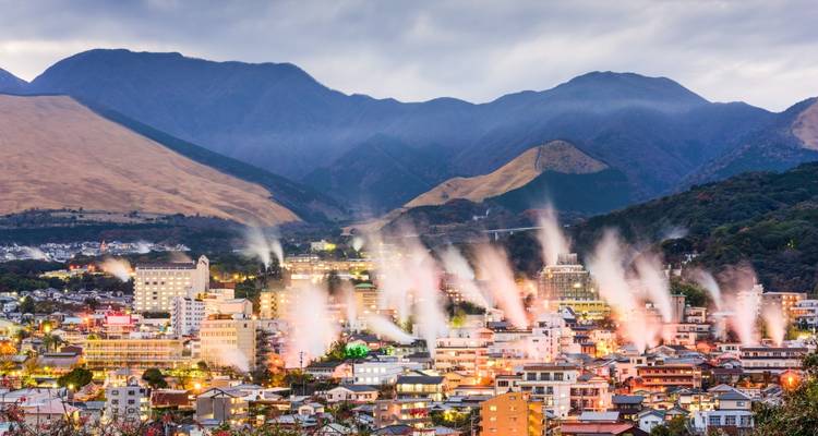 Panorama du soir d'une ville avec de nombreux panaches de vapeur s'élevant contre un arrière-plan de montagnes sombres.