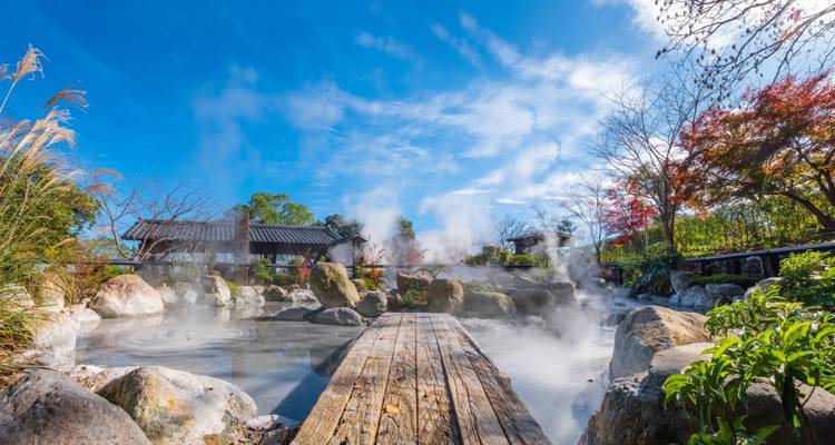 Vapeur s'élevant d'un bassin d'onsen rocheux extérieur avec une passerelle en planches de bois sous un ciel bleu éclatant.
