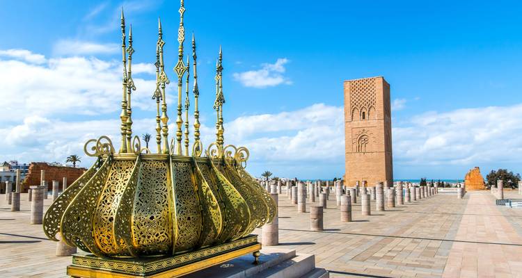 Monumento dorado ornamentado en una plaza abierta frente a la Torre Hassan bajo un cielo azul brillante.