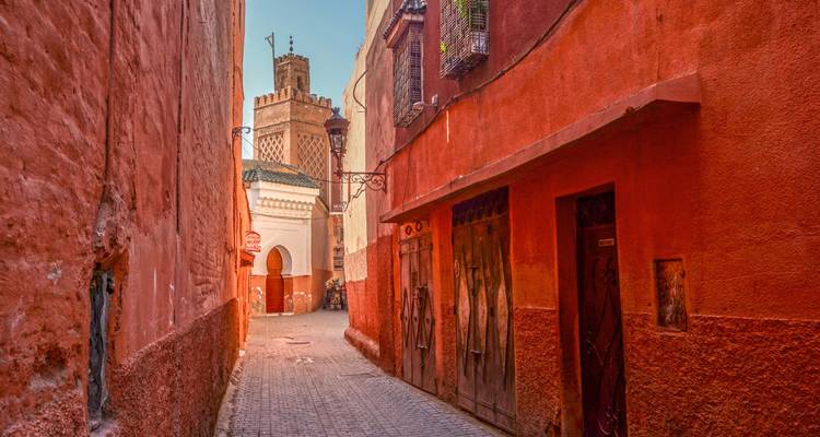 Callejón estrecho de paredes rojas en una medina antigua que conduce hacia un minarete distante.