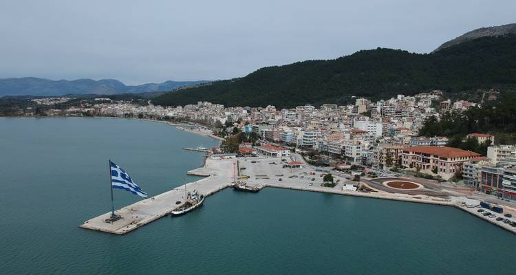 Luchtfoto van de haven van Igoumenitsa met een enorme Griekse vlag en een stad aan het water met groene heuvels op de achtergrond.