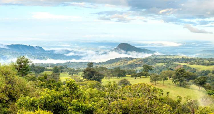 Vaste panorama de collines vertes ondulantes et de vallées avec des nuages dérivant sous un sommet lointain.
