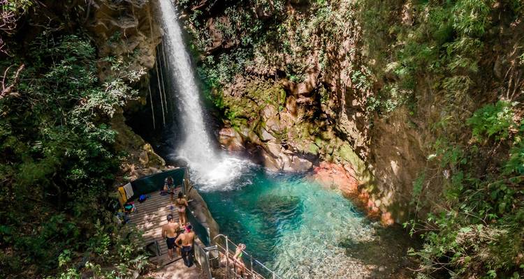 Les nageurs profitent d'un bassin de cascade turquoise accessible par une terrasse et des escaliers en bois.