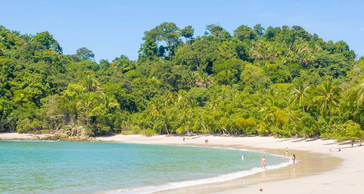 Visitors wade and sunbathe on a curved white-sand beach edged by jungle.