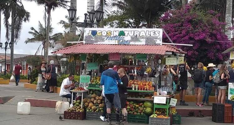 Petit stand de jus à ciel ouvert sur une place de ville avec des fruits tropicaux exposés et plusieurs touristes commandant des boissons