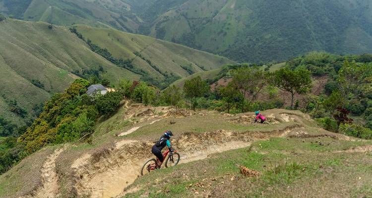 Deux cyclistes descendent un sentier de terre sinueux sur un flanc de montagne verdoyant avec des vues panoramiques sur la vallée
