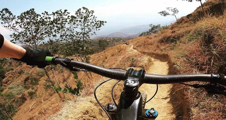 Point de vue du guidon le long d'un étroit sentier de montagne poussiéreux bordé d'herbe sèche et d'arbres épars