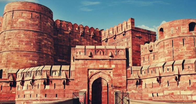 Massive geschwungene Bastionen aus rotem Sandstein und das verzierte Tor des Agra-Forts erheben sich unter einem leuchtend blauen Himmel.