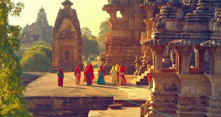Frauen in leuchtenden Saris gehen durch sonnenbeschienene Sandstein-Tempelgänge mit üppigem Grün in der Ferne.
