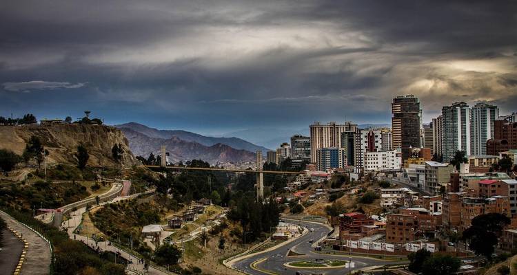 Vista amplia de La Paz con un puente que atraviesa un valle profundo, rascacielos modernos y picos andinos bajo un cielo dramático nublado.