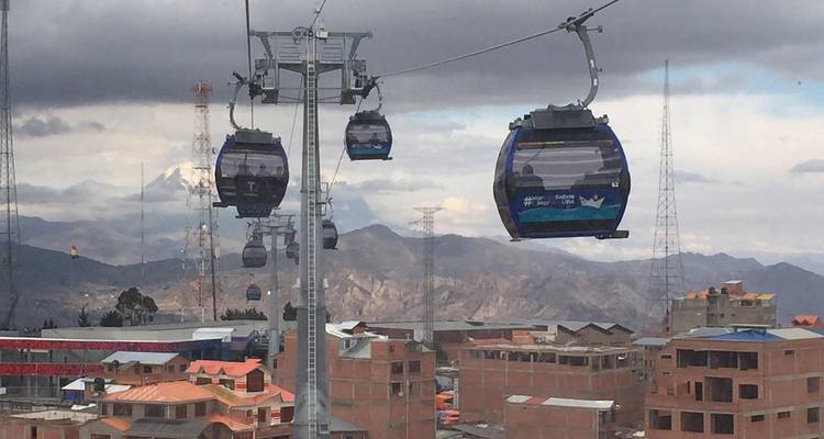 Múltiples cabinas azules del Mi Teleférico viajan por encima de edificios de ladrillo con picos andinos nevados al fondo.