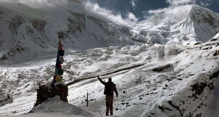 Een eenzame trekker steekt een arm op naar torenhoge gletsjerwanden onder dramatische wolken.
