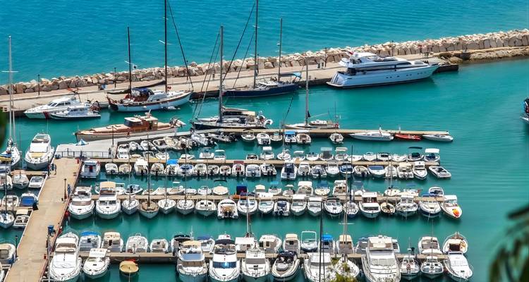 Des rangées de yachts et de bateaux sont amarrés dans une marina turquoise protégée par une digue en pierre.